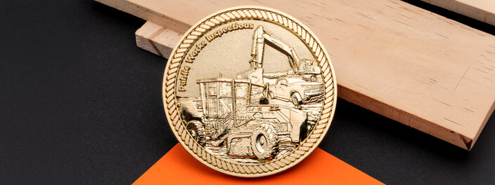 Round gold coin with a sculpted excavator, dump truck and grader inside a rope border, on light wood