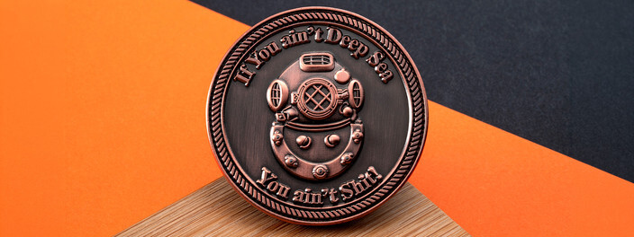 Round copper coin with a sculpted deep sea diving helmet inside a rope border, on orange paper