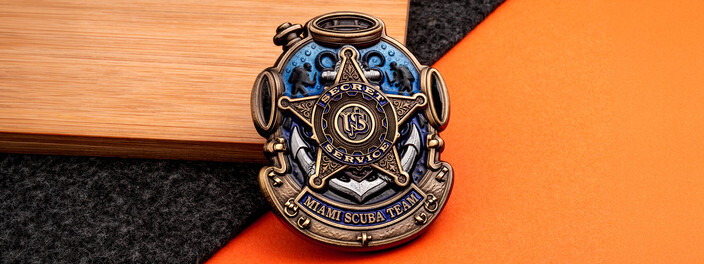 Diving helmet shaped coin with a bronze star badge over an anchor and two divers in shaded blue water, on wood, gray felt and orange paper