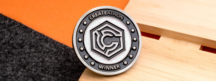Silver Createathon winner coin with a hexagon logo on white glitter enamel, on light wood and orange paper