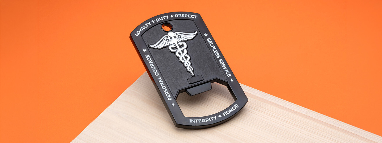 Black dog tag shaped bottle opener coin with a white caduceus and an Army values motto around the edge, on light wood and orange paper