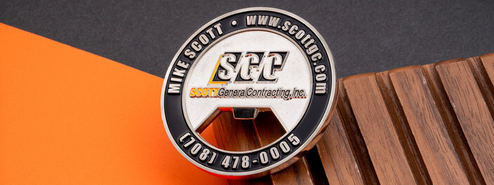 Round silver bottle opener coin with a contracting company's logo, website and phone number, on dark wood
