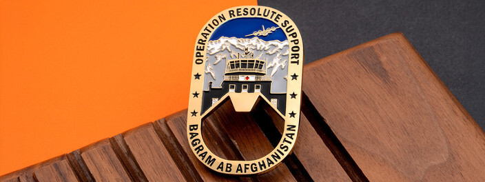 Gold oval bottle opener coin with an air base control tower under snowy mountains, on dark wood and orange paper