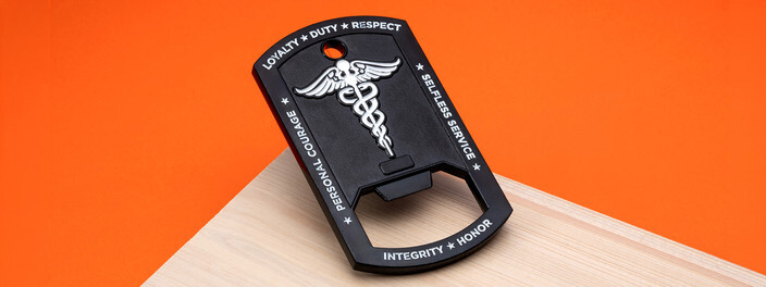 Black dog tag bottle opener with a silver caduceus and a ring of engraved values, on pale wood against orange
