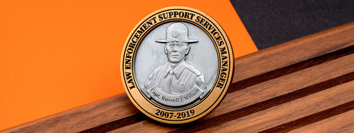 Gold rimmed coin with a silver portrait of a captain in a campaign hat and the years 2007 to 2019, on dark wood