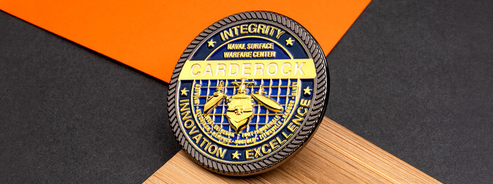 Navy and gold naval surface warfare center coin with a gold ship over a blue grid, on light wood and orange paper