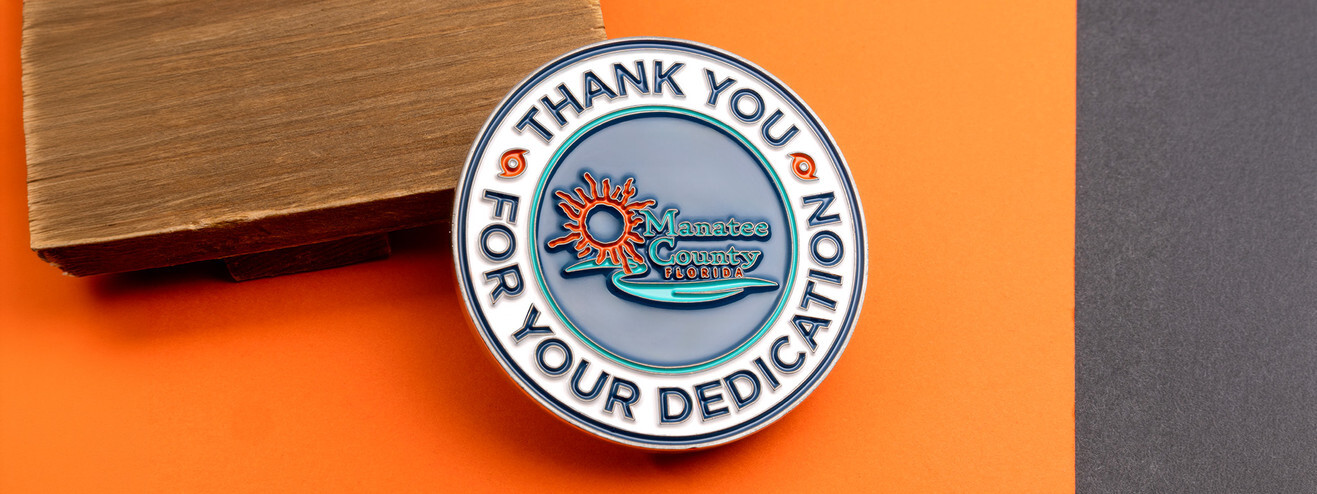 Silver county coin reading Thank You for Your Dedication around a sun and water logo, on wood and orange paper