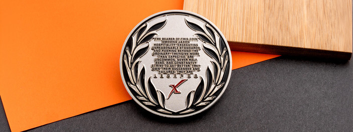 Silver coin with a black laurel wreath around a company pledge that ends with the word Legends, on orange paper and wood