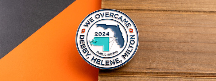 Public works coin reading We Overcame Debby, Helene, Milton around a 2024 Florida map, on wood and orange paper