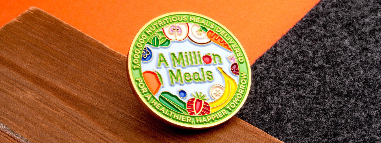 A Million Meals coin with a green rim and fruit and vegetables in color enamel, on dark wood and orange paper