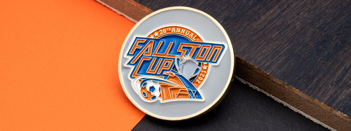 Gray soccer tournament coin with orange and blue lettering, a ball and a trophy