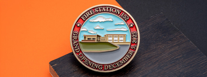 Gold fire station grand opening coin with an enamel scene of the new station under a blue sky, on dark wood