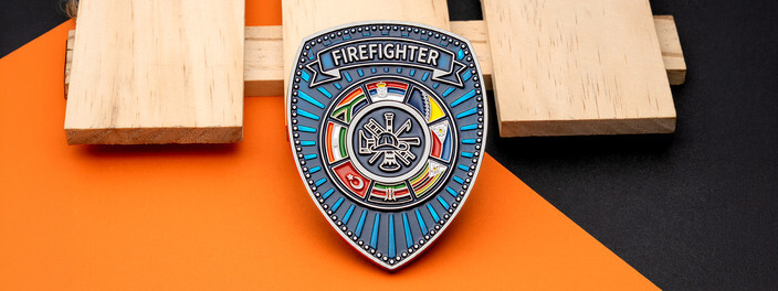 Shield shaped firefighter coin with a ring of national flags around a fire department emblem, on light wood and orange paper